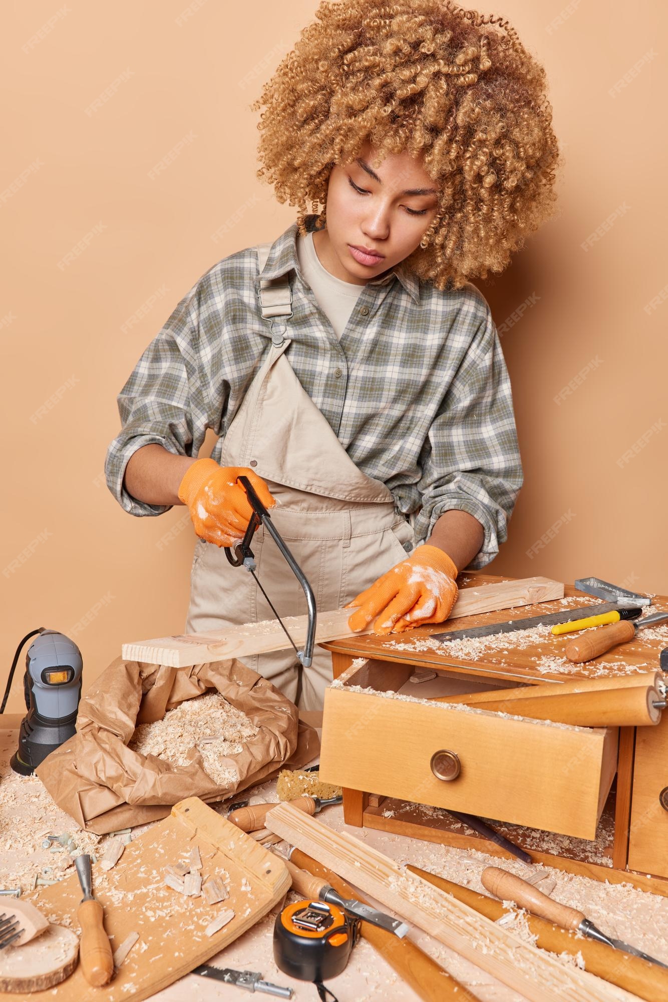 Tiro vertical de carpinteiro profissional feminino trabalha na oficina  corta madeira com quebra-cabeças faz móveis vestidos com poses de roupas de  trabalho contra a parede marrom com instrumentos ao redor mestre qualificado