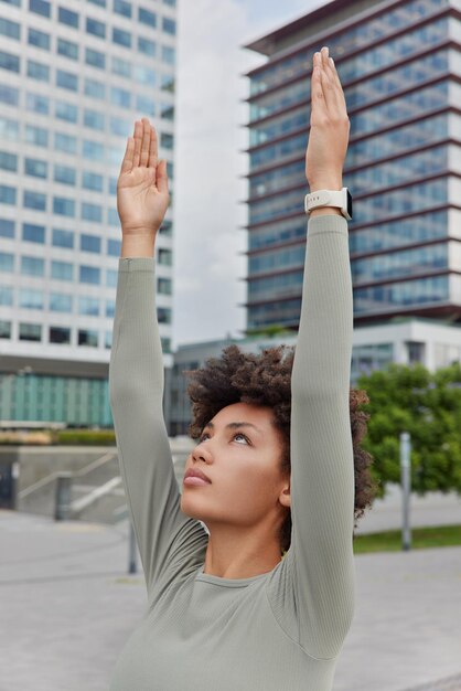 Foto tiro vertical de esportista de cabelos encaracolados vestido com roupa ativa levanta os braços focados acima de exercícios ao ar livre em ambiente urbano faz exercícios regulares durante a manhã conceito de estilo de vida saudável