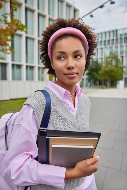 Tiro vertical de estudante pensativa caminha para a universidade carrega livros e poses de tablet com mochila usa roupas legais poses ao ar livre contra o meio urbano. educação e estudo