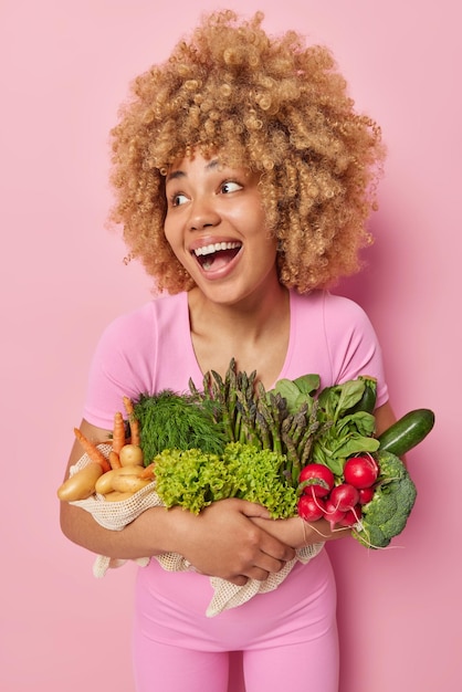 Tiro vertical de mulher de cabelo encaracolado feliz abraça um monte de legumes frescos vestidos com roupas casuais focados longe mantém a dieta saudável isolada sobre fundo rosa mercearia verde do próprio jardim