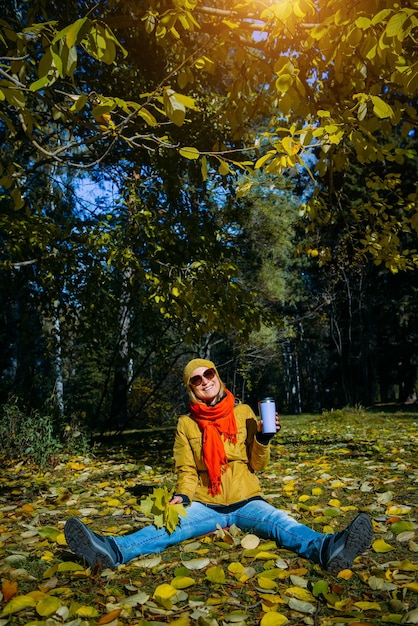 Tiro vertical de mulher feliz, aproveitando o belo dia de sol no parque outono. Jovem com copo térmico e folhas de plátano amarelas nas mãos dela sentada no chão coberto com folhagem caindo.