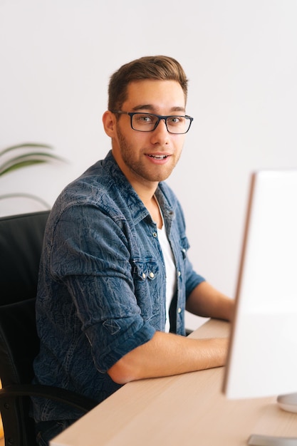 Tiro vertical do macho programador freelance jovem confiante em óculos elegantes, trabalhando no computador desktop, sentado na mesa no escritório em casa, na sala com interior moderno, olhando para a câmera.