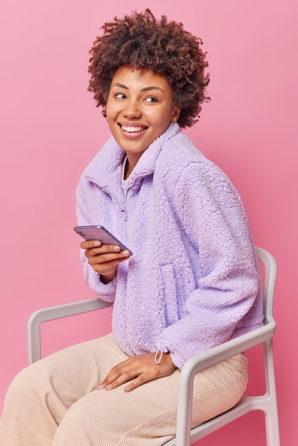 Tiro vertical de feliz mujer de pelo rizado vestida con abrigo y pantalones se sienta en una silla utiliza chats de teléfono móvil en línea espera llamada aislada sobre pared rosa