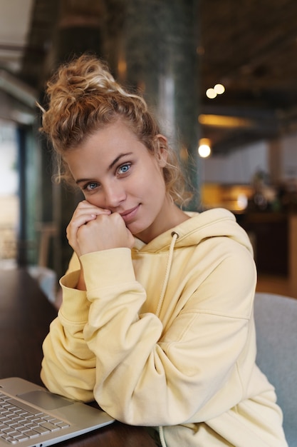 Tiro vertical de hermosa niña sonriente sentada en un café con laptop, escritorio de mesa inclinada y mirando la cámara.