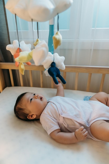 tiro vertical inocente bebê asiático deitado no berço em babysuit está olhando para o brinquedo móvel sobre sua cabeça curiosamente enquanto brincava sozinho durante a hora de dormir no quarto.
