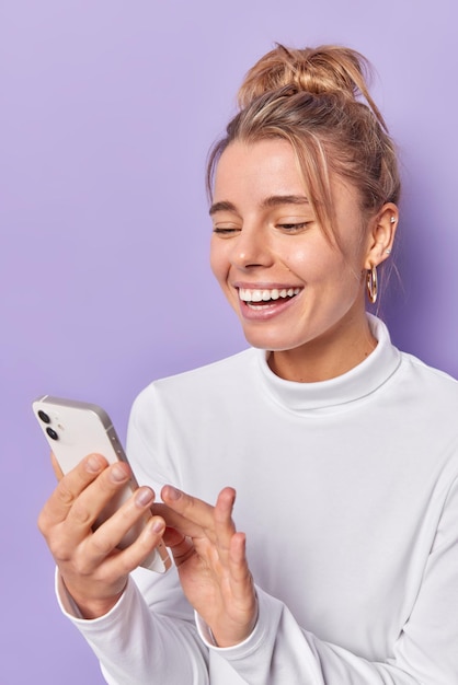 Tiro vertical de mujer alegre y feliz revisa el suministro de noticias a través de sonrisas de teléfonos inteligentes, disfruta ampliamente de la comunicación en línea y usa un jersey blanco informal aislado sobre fondo púrpura. Concepto de tecnología.