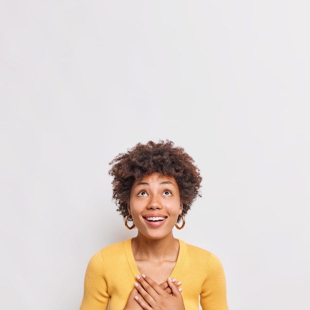 Tiro vertical de mujer joven feliz con cabello rizado presiona las manos en el pecho expresa gratitud centrada arriba tiene una sonrisa con dientes viste un suéter amarillo aislado sobre una pared blanca