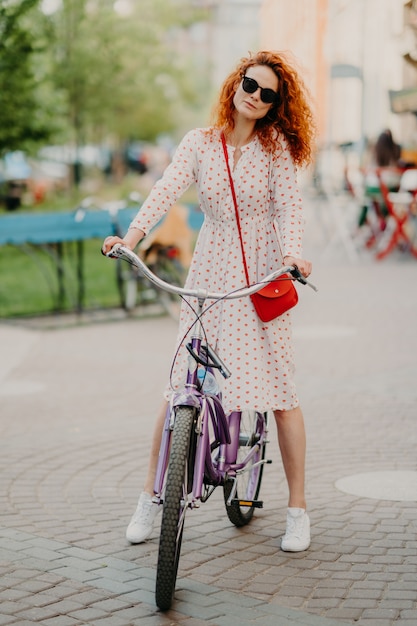 Tiro vertical de mujer pelirroja rizada monta bicicleta en la ciudad durante la semana, tiene tiempo libre