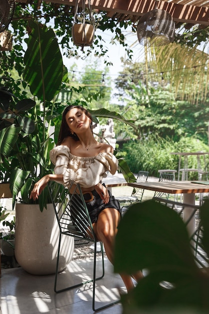 Foto tiro vertical de una mujer sensual sentada en un café rodeado de hojas verdes, palmeras y plantas tropicales.
