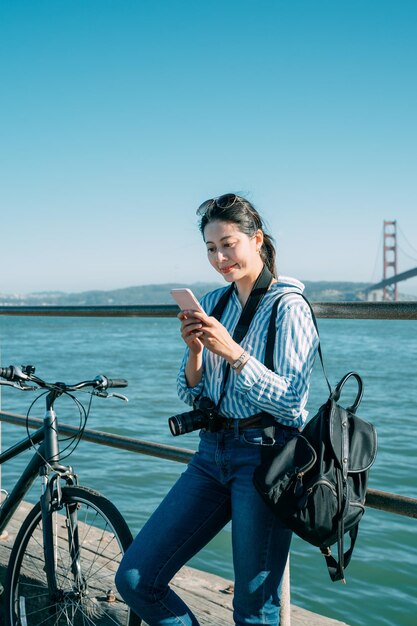 tiro vertical na moda menina asiática em pé junto ao parapeito à beira-mar com uma bicicleta está desfrutando de feeds de mídia social no smartphone sob o céu azul ensolarado na Califórnia.