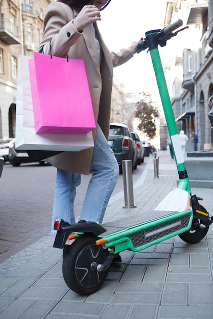 Tiro vertical recortada de mujer irreconocible con bolsas de compras pisando scooter eléctrico en la calle