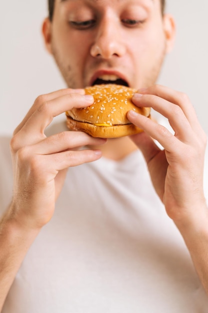 Tiro vertical recortado de un joven apuesto que disfruta comiendo una deliciosa hamburguesa en un fondo blanco aislado. Retrato de primer plano de un hombre feliz y guapo sosteniendo una sabrosa hamburguesa malsana.