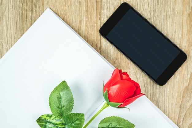 Tiro de vista superior de rosas rojas en diario de cuaderno en blanco y teléfono inteligente en mesa de madera, concepto de San Valentín