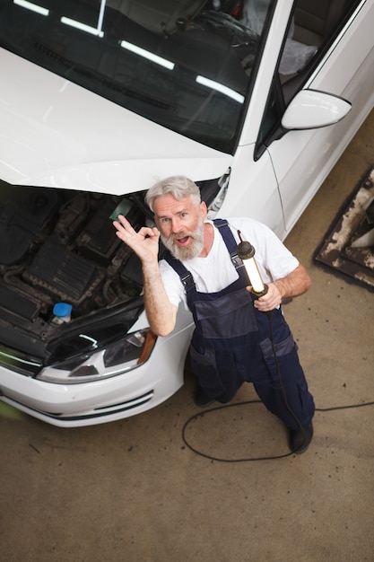 Tiro de vista superior vertical de un reparador de servicio de coche anciano alegre que muestra el signo de ok a la cámara