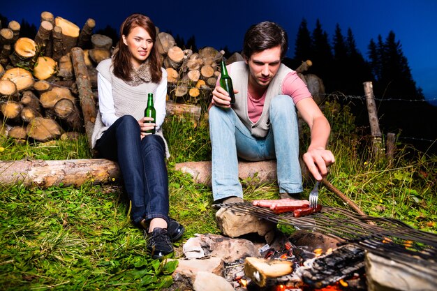 Tirol - Junges Paar sitzt auf der Almwiese eines Berges am Lagerfeuer in den bayerischen Alpen und genießt den romantischen Abendsonnenuntergang des Panoramas in der Freizeit oder im Urlaub