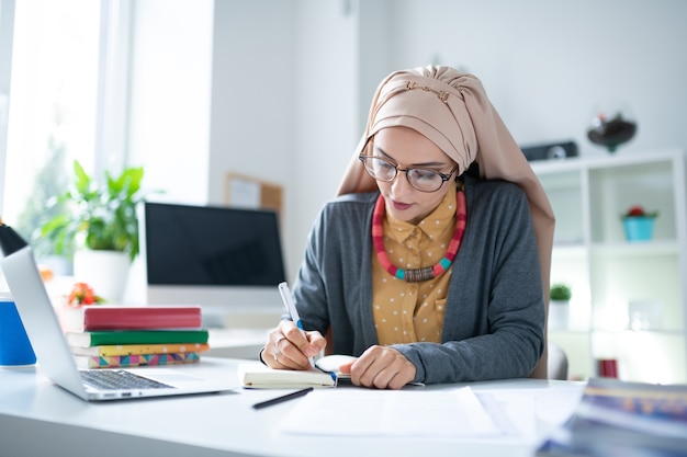 Tisch mit Büchern. Beschäftigter muslimischer Lehrer mit Hijab, der mit Büchern am Tisch sitzt und arbeitet