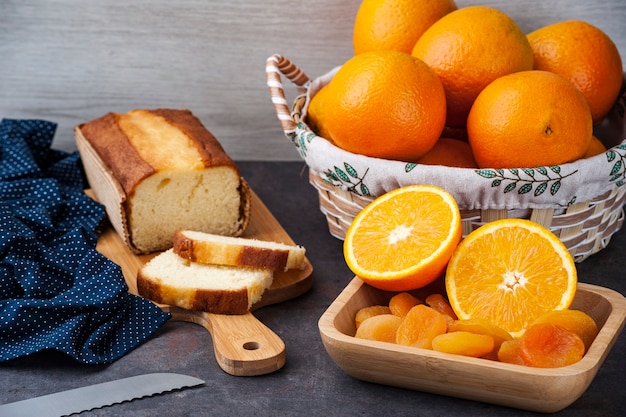 Tisch mit ganzen und geschnittenen orangen, getrockneten aprikosen und orangenkuchen auf einem küchenbrett.