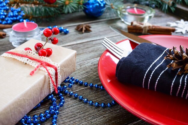 Tischdekoration zu Weihnachten. Rote Platte, Gabel, Messer, Kerze, Serviette, Geschenkzweig eines Weihnachtsbaumes auf einem hölzernen Hintergrund. Weihnachten Weihnachten Neujahr Urlaub Postkarte.