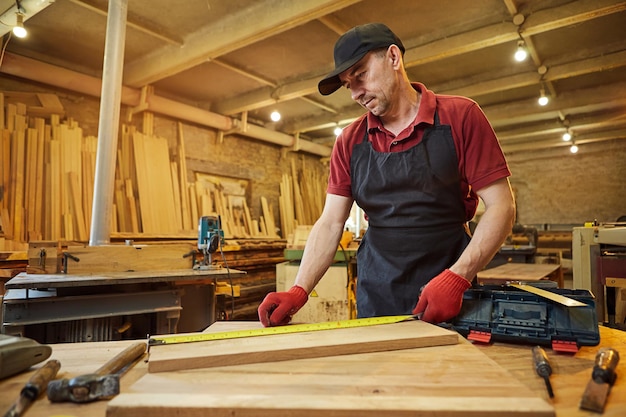 Tischler, der mit einem Bleistift mit einer Holzmarkierungsplanke arbeitet und Messungen vornimmt, um ein Stück Holz zu schneiden, um ein Möbelstück in einer Tischlerei herzustellen