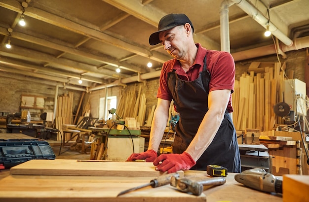 Tischler, der mit einem Bleistift mit einer Holzmarkierungsplanke arbeitet und Messungen vornimmt, um ein Stück Holz zu schneiden, um ein Möbelstück in einer Tischlerei herzustellen