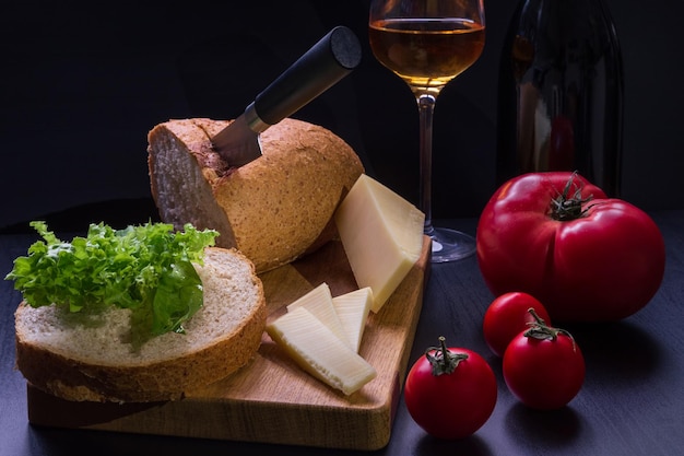 Tischplattenstillleben mit Brotkräutern, Tomaten, Käse, Weinglas und dunkler Flasche