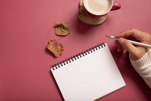 Tischschreibtisch mit Notizblock und weiblicher Hand mit Bleistift und Herbstblättern auf burgunderfarbenem Hintergrund Herbstliche flache Lay-Komposition