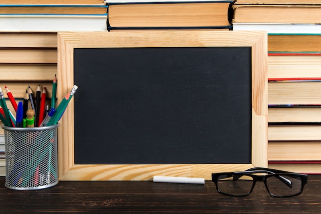 Tiza pizarra, gafas, soporte con bolígrafos, lápices y tizas, contra libros, copia espacio.