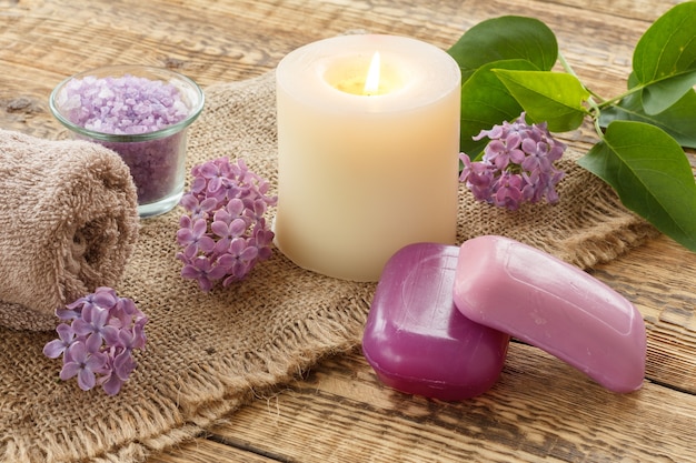 Toalla de felpa, jabón para procedimientos de baño, sal marina, vela encendida y flores lilas sobre cilicio y viejas tablas de madera. Productos y accesorios de spa.