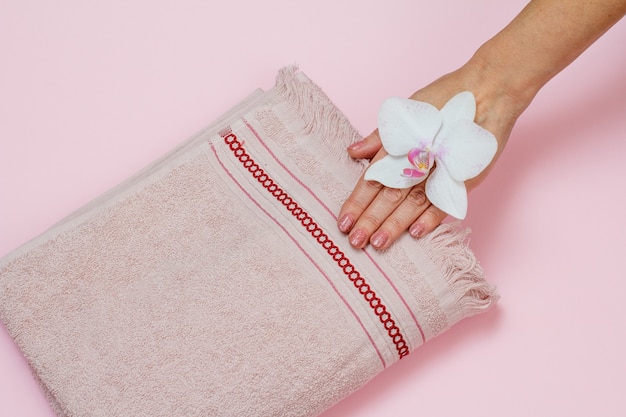 Toalla de felpa suave, mano de mujer con flor de orquídea blanca sobre fondo rosa. Vista superior.