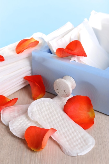 Toallas sanitarias en caja y pétalos de rosa sobre mesa de madera sobre fondo azul claro