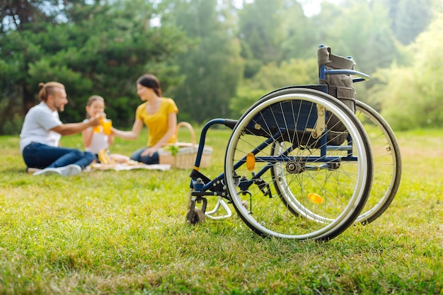 Toast auf die Gesundheit. Der Fokus liegt auf einem Rollstuhl eines angenehmen jungen Mannes, der mit seiner Familie bei einem Picknick im Wald Gläser mit Saft anstößt