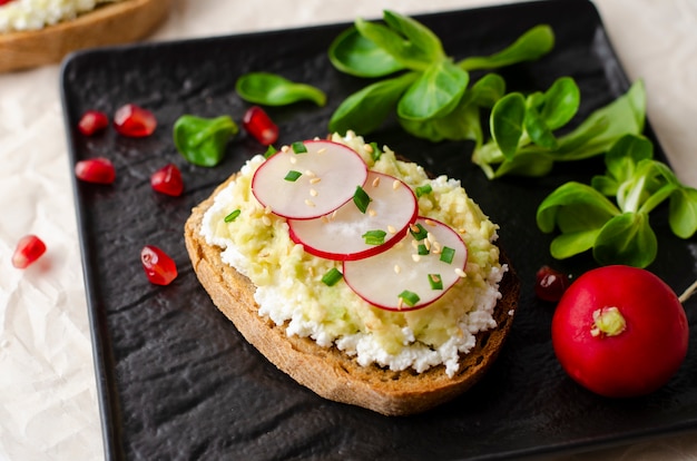 Toast mit Hüttenkäse und zertrümmerter Avocado, Rettich, Feldsalat und Granatapfelkernen auf Schwarzblech.