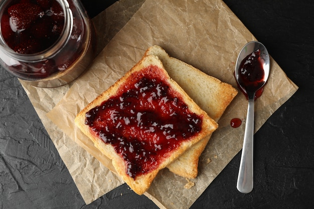 Toast und Glas mit Marmelade, Löffel und Backpapier auf schwarzem Hintergrund, Draufsicht