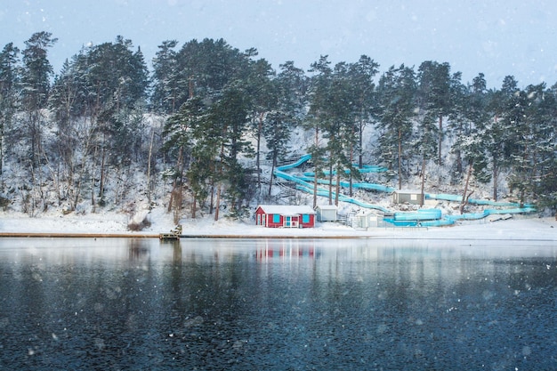 Tobogán de agua vacío con cabaña de playa roja cerca del lago congelado en un frío día de invierno con nieve en las nubes. Suecia