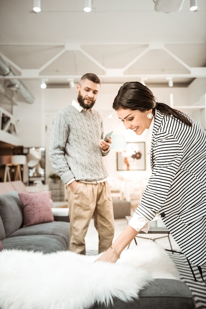 Tocando pelaje esponjoso. Señora positiva sonriente que muestra una decoración esponjosa a su esposo mientras él está de pie con el teléfono inteligente