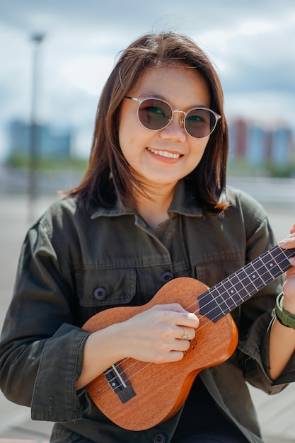 Tocando el ukelele de la joven y bella mujer asiática vistiendo chaqueta y jeans negros posando al aire libre