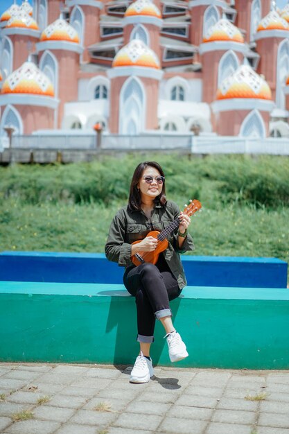 Tocando el ukelele de la joven y bella mujer asiática vistiendo chaqueta y jeans negros posando al aire libre