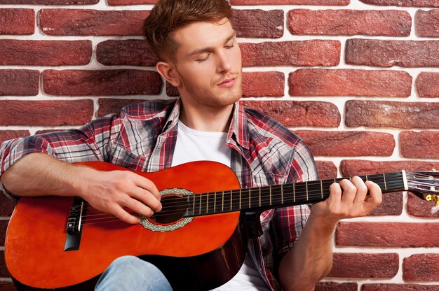 Tocando violão. Jovem bonito tocando violão enquanto se inclina para a parede de tijolos