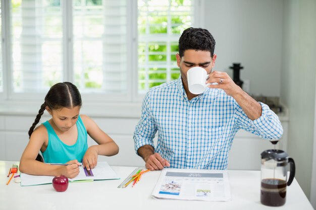 Tochter macht ihre Hausaufgaben und Vater trinkt eine Tasse schwarzen Tee beim Lesen der Zeitung