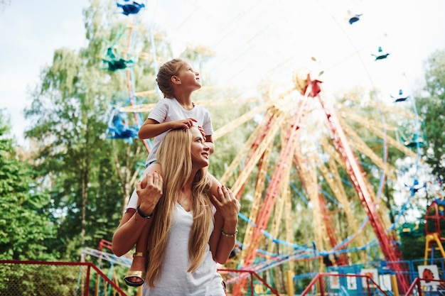 Tochter sitzt auf den Schultern. Fröhliches kleines Mädchen, ihre Mutter, haben eine gute Zeit im Park zusammen in der Nähe von Attraktionen.