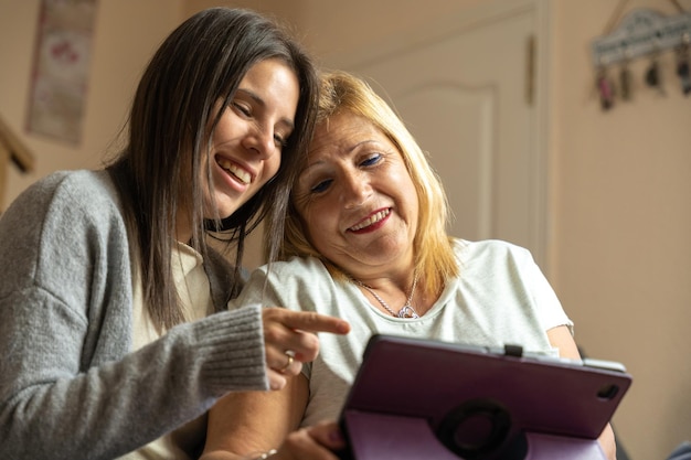 Tochter zeigt ihrer Mutter etwas auf dem Tablet und die Mutter ist aufmerksam, während sie zu Hause glücklich auf dem Sofa sitzen