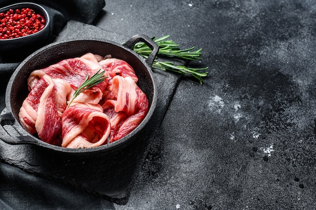 Tocino crudo, tiras de carne veteada en una sartén. Fondo negro. Vista superior. Copie el espacio.