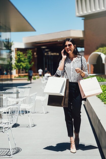 toda la longitud de una hermosa dama asiática elegante hablando por teléfono celular sonriendo alegre. niña cargando muchas bolsas de compras comprando en la tienda de ropa en el centro comercial de stanford. mujer charlando al aire libre de pie