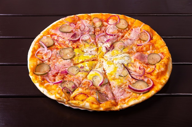 Toda una pizza rústica con pepino encurtido, tocino, aros de cebolla y huevos fritos se sirve en un plato de papel sobre una mesa de madera