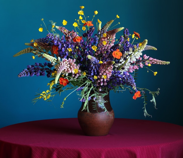 Todavía vida con un ramo del lupine y de ranúnculos del verano en el jarro en la mesa redonda con un mantel rojo en un fondo azul.