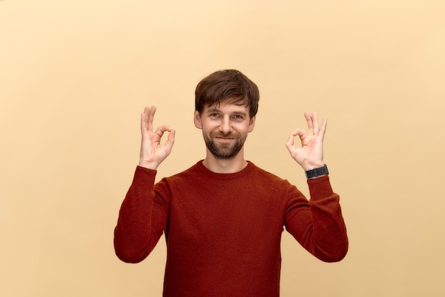 Foto todo está bien. foto de hombre joven con barba con suéter, muestra signos de bien, tiene una sonrisa, posando contra la pared beige.