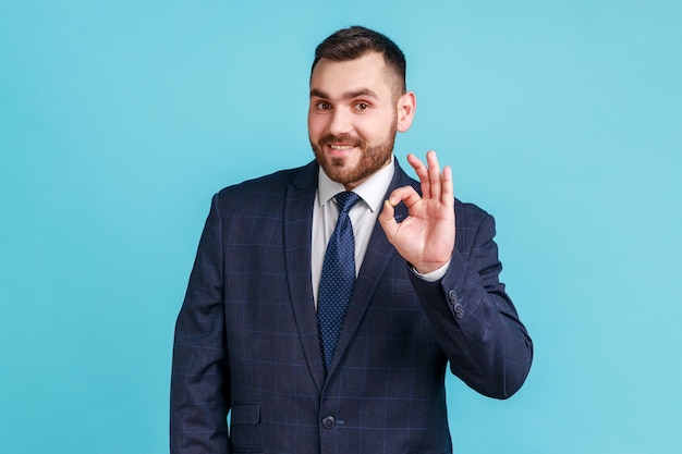 Todo esta bien. Hombre barbudo sonriente con traje de estilo oficial, mirando la cámara natural, mostrando el signo correcto, expresando emociones positivas. Disparo de estudio interior aislado sobre fondo azul.