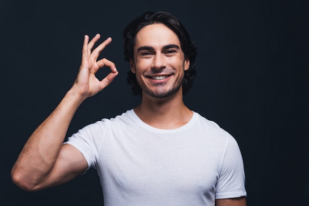 Foto todo está bien. hombre joven feliz gesticulando el signo de ok y mirando a la cámara mientras está de pie