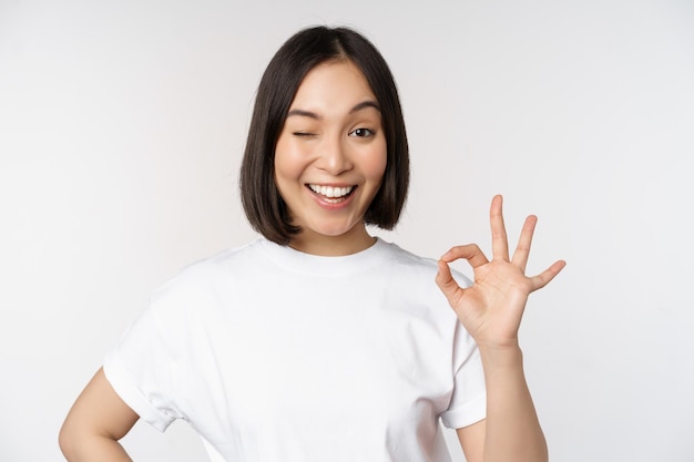 Todo bien Sonriente joven asiática asegurando que muestra el signo de ok con la cara satisfecha de pie sobre fondo blanco.