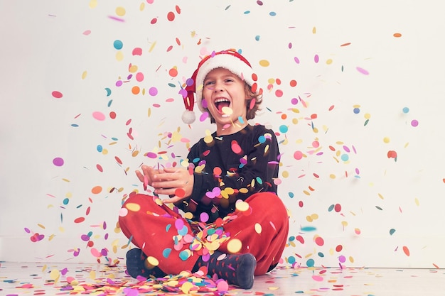 Todo el cuerpo de un adolescente encantado con sombrero de Papá Noel mirando hacia arriba mientras se sienta bajo confeti brillante que cae sobre fondo blanco durante la celebración de Navidad